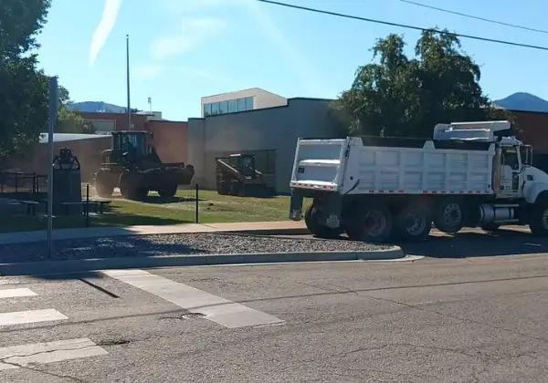 Dump truck and front loader working together on a street corner near a school building