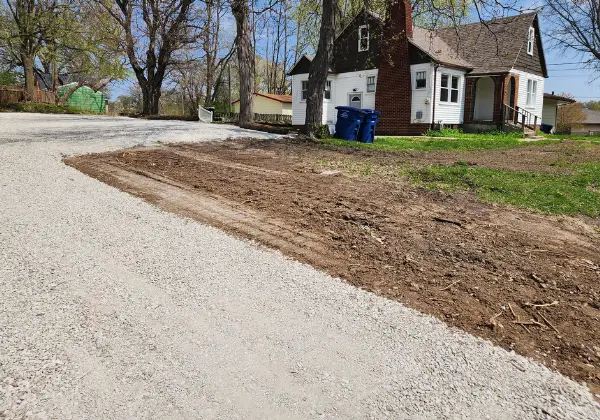 Freshly graded bare dirt strip along a gravel driveway next to a brick house with two trash bins