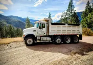 White Maxx Transfer dump truck parked on a gravel road with forested mountains in the background