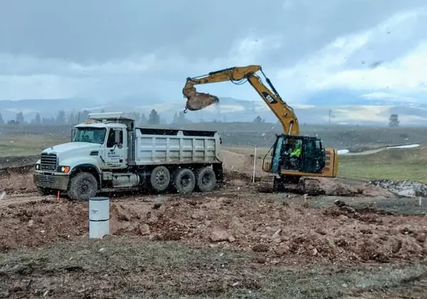 Excavator loading dirt into a Maxx Transfer dump truck on an overcast day in an open field